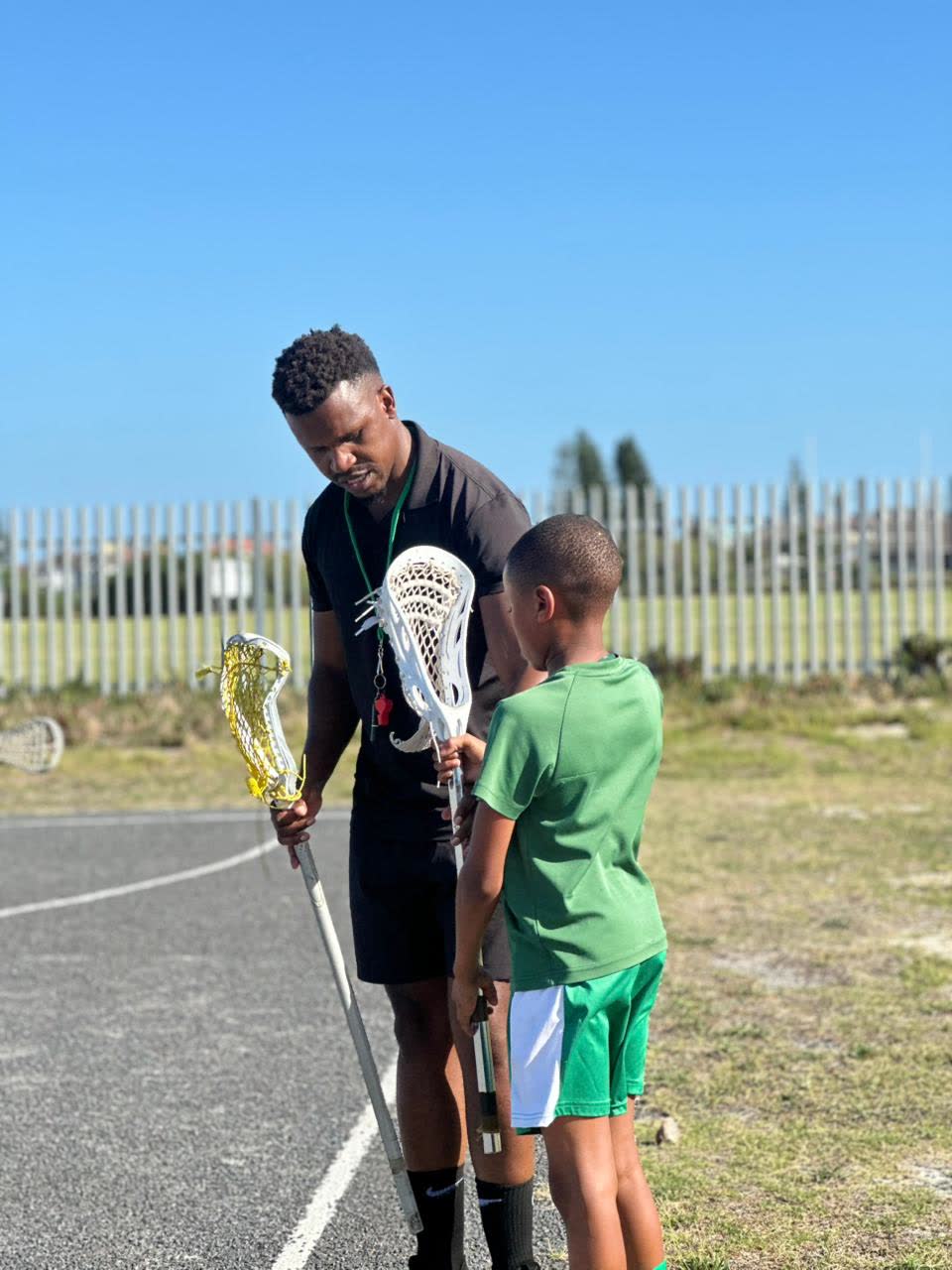 A coach handing a lacrosse stick to a young player in Muizenberg