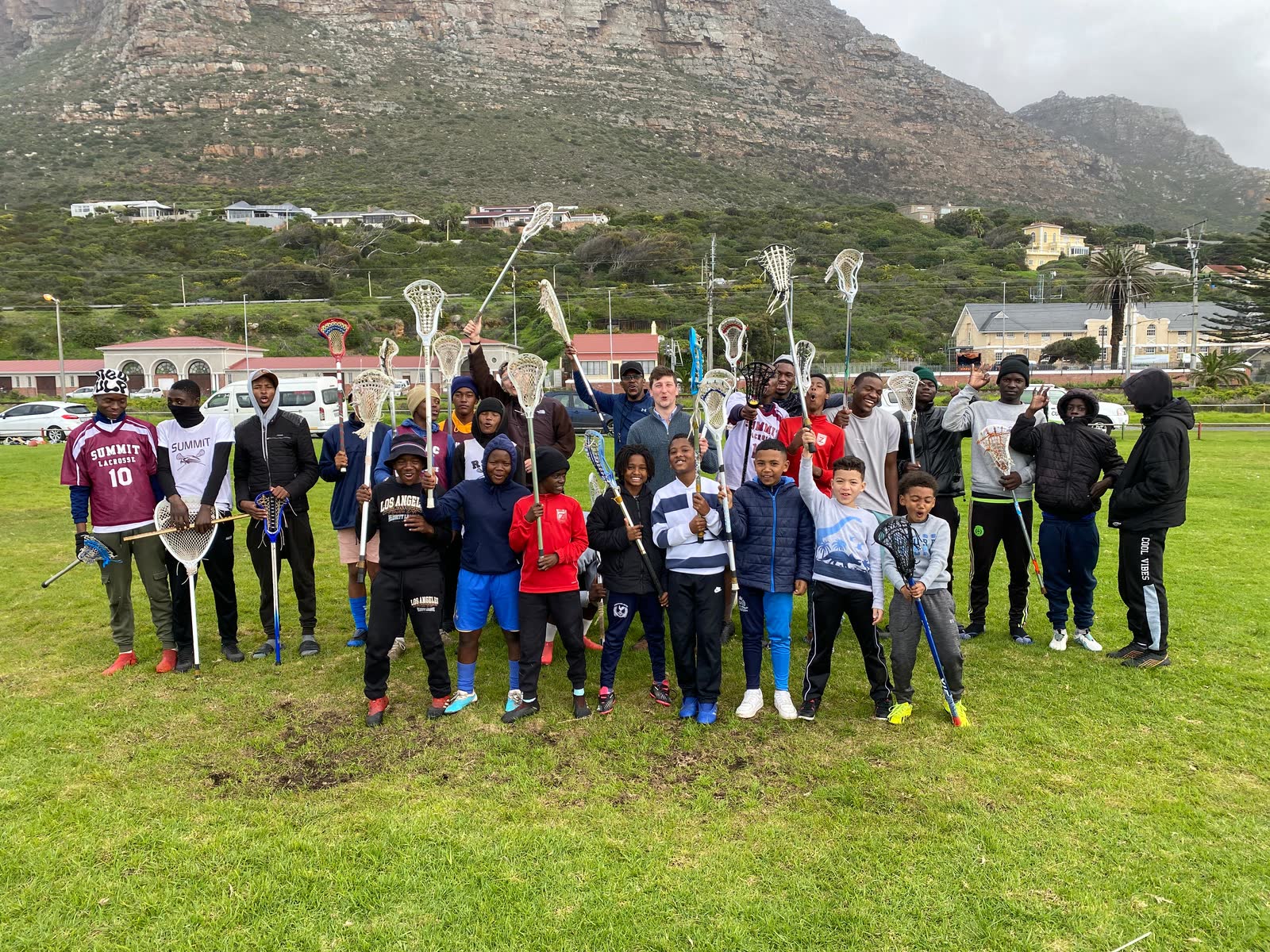 A large group of players and coaches with sticks raised in Muizenberg, mountains in the background